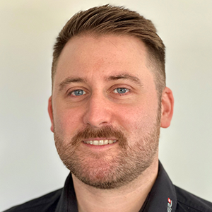 Ein Mann mit kurzem, hellbraunem Haar und Bart laechelt in schwarzem Hemd vor hellem Hintergrund, Fachmann Blechbearbeitung.