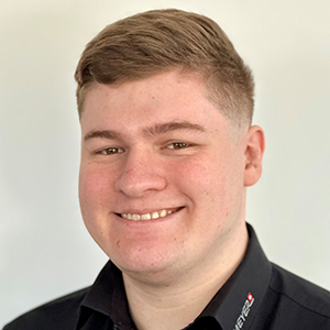 Junger Mann mit kurzem, hellbraunem Haar lacht in die Kamera, trägt schwarze Bluse vor hellem Hintergrund, Blechbearbeitung.