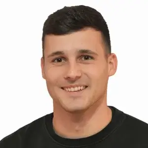Ein junger Mann mit kurzen dunklen Haaren, im schwarzen Shirt, lacht vor weißem Hintergrund wie in einer Blechbearbeitungsfirma.