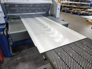 Ein Blech läuft aus einer industriellen Walzanlage, im Hintergrund sind Paletten und Maschinen zu sehen; sichtbare Spuren der Blechbearbeitung.