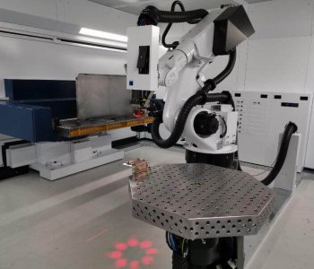 Ein Roboterarm mit Greifer steht in einem modernen Industrie-Labor neben Blechbearbeitungs-Tisch und Steuerpulten.