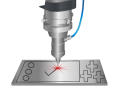 Eine Lasergravurmaschine markiert ein Häkchen auf einer blechartigen Oberfläche mit Controller-Tasten und Steuerkreuz.