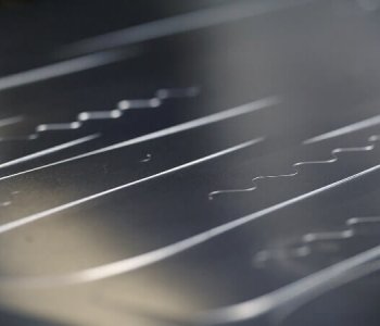 Nahaufnahme einer Metalloberfläche mit geraden und welligen Prägungen, Lichtreflexen und feinen Schatten im Blech.