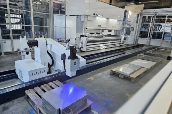 Moderne automatisierte Blechbearbeitungsmaschine, Metallplatten auf Holzpalette, Glaswände und hohe Decken in Fabrikumgebung.