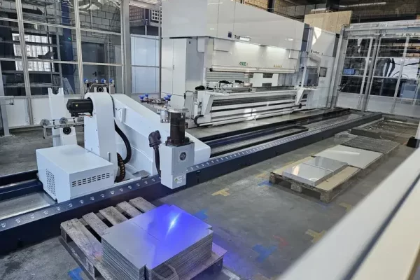 Moderne automatisierte Blechbearbeitungsmaschine, Metallplatten auf Holzpalette, Glaswände und hohe Decken in Fabrikumgebung.