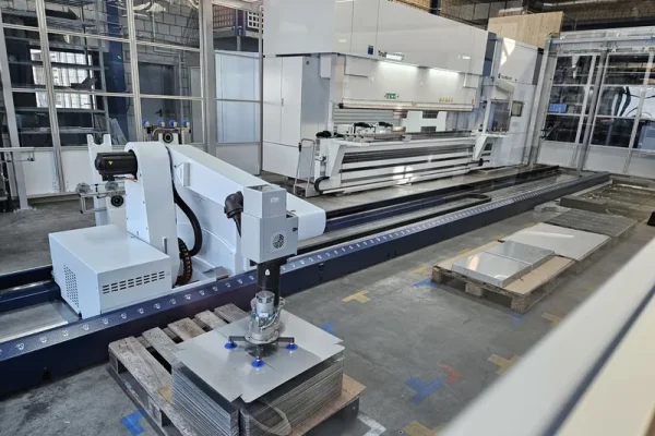 Ein Roboterarm mit Saugheber hebt Bleche vom Palettenstapel in einer Industriehalle mit Maschinen und Glastrennwänden.