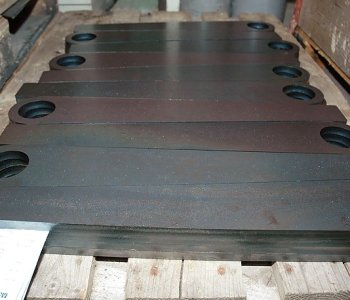 Mehrere große, flache Blechteile mit runden Enden und Lochung liegen ordentlich auf einer Holzpalette; eine weisse Notiz sichtbar.