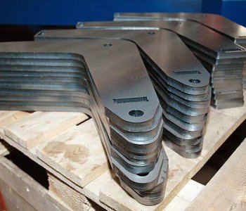 Ein Stapel identischer, L-förmiger Metallwinkel mit abgerundeten Kanten liegt auf einer Holzpalette in einer Blechbearbeitungshalle.