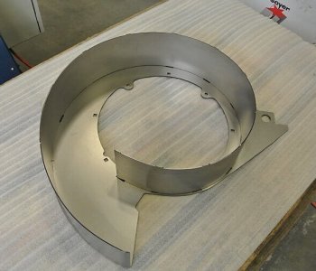 Ein spiralförmiges Metall-Blechteil mit Loch liegt auf Wellblech im Werkstattumfeld, halbgeschlossen und gekruemmt.