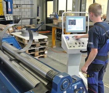 Ein Arbeiter im blauen Overall bedient ein Steuerpult an einer Walzmaschine, Bleche und Paletten sind im Hintergrund sichtbar.