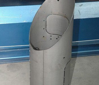 Ein zylindrisches Blech mit schräger, ovaler Öffnung und Bohrungen steht auf Beton vor einer metallisch-blauen Oberfläche.