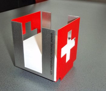 Metall-Serviettenhalter mit ausgeschnittetem Schweizer Kreuz, rot lackiert, auf grauem Tisch, Gravur "www.laserschneiden.ch".