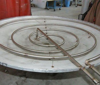 Große Metallspirale auf runder Platte in Werkstatt für Blechbearbeitung, zwei Rohre führen seitlich weg, Werkzeug im Hintergrund.