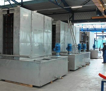 Grosse industrielle Metall-Kühltürme mit blauen Motoren stehen in einer Halle, daneben zwei Arbeiter und Regalreihen mit Ausrüstung.
