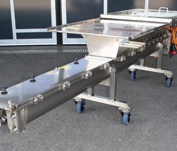 Edelstahl-Förderband auf Rollen mit transparenter Abdeckung und Steuerung draussen, orange Elektrokabel rechts sichtbar.