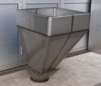 Großer, industrieller Edelstahltrichter mit quadratischem Einlass und schmalem Auslass auf Betonboden bei Metallwand.