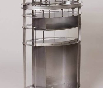 Zylindrischer Metallrahmen mit zwei Tablaren, offenen Seiten und industriellem Look aus Blechbearbeitung, minimalistisch gefertigt.
