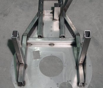 Ein Metallrahmen mit grober gelochter Rundbasis und mehreren Stützblechen steht auf grauem Betonboden.