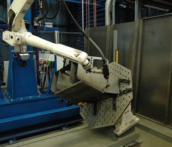 Ein Roboterarm schweißt ein Metallbauteil auf eine Lochvorrichtung, umgeben von Maschinen, Kabeln und Blechpaneelen.