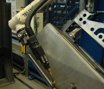 Ein Roboterarm mit Schweisswerkzeugen bearbeitet ein Blechteil in einer Industriehalle mit blauen Stahlträgern und Maschinen.