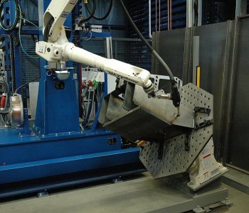 Ein Roboterarm bearbeitet ein grobes Blechteil auf einem Lochwerktisch. Blaue Maschinen und Schutzbarrieren im Hintergrund.
