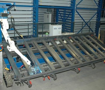 Ein Industrieroboter schweisst an einem großen Metallrahmen, fixiert mit Klemmen, vor blauen Regalen in der Werkhalle.