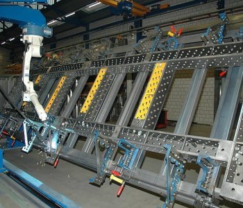 Ein Industrieroboter schweisst eine grosse, rechteckige Stahlbaugruppe in eine Werkstatt für Blechbearbeitung, fixiert mit Spannvorrichtungen.