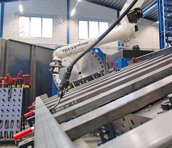 Ein Schweissroboter bearbeitet Metalltraeger mit Spannvorrichtungen in einer Werkstatt, umgeben von Maschinen und Regalen.